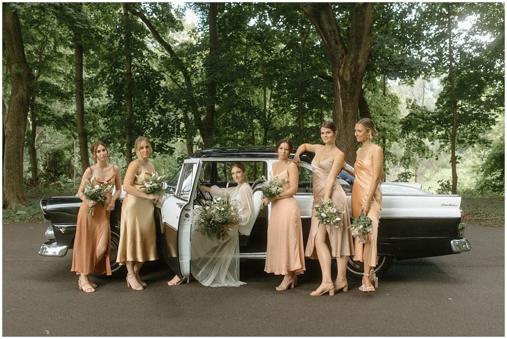 Sarah's bridesmaids stand on either side of her in front of a vintage car at The Cooperage.