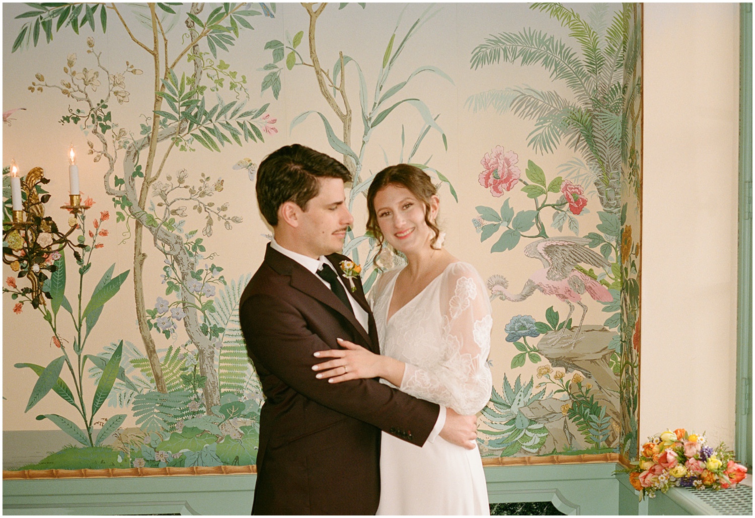 A groom puts his arms around a bride's waist in front of a wall of floral wallpaper at Villa Terrace Milwaukee.