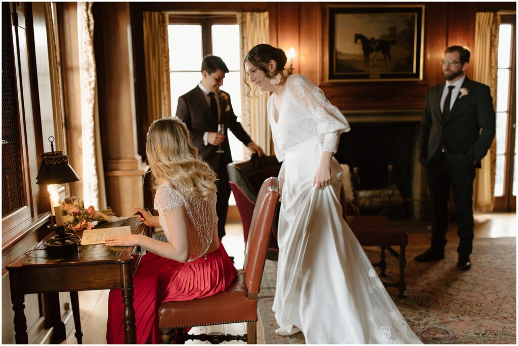 A bride looks over the shoulder of a bridesmaid sitting and signing a marriage license.