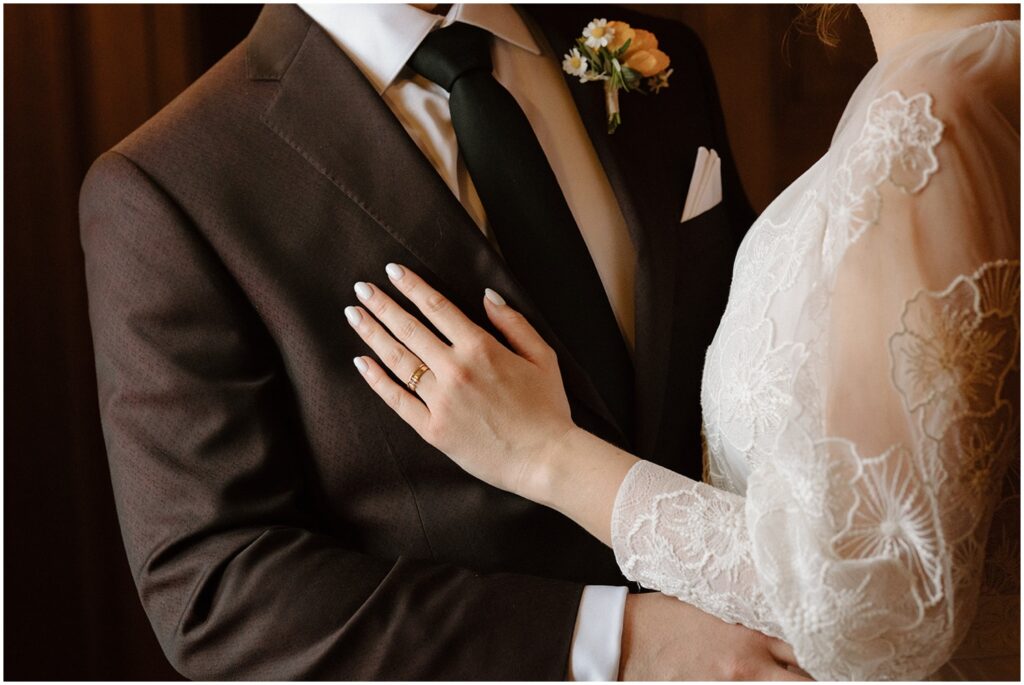 A bride rests a hand on a groom's chest, showing her wedding ring.