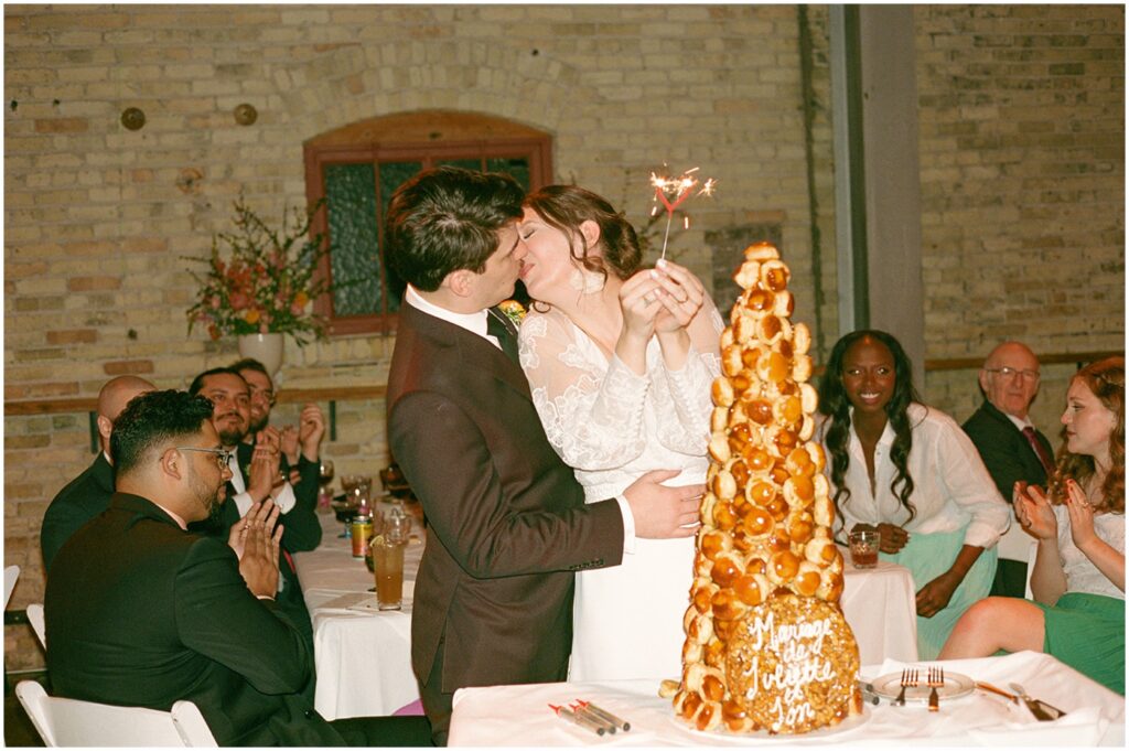 A bride and groom put a sparkler on a tower of puff pastries.