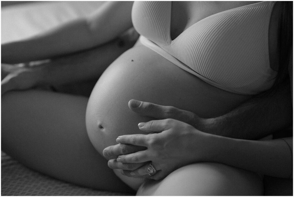 A man sits behind a woman and holds her hand against a baby bump in a lifestyle maternity photo.