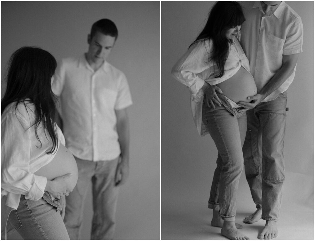A man stands behind his pregnant wife looking at her baby bump in a studio maternity portrait.