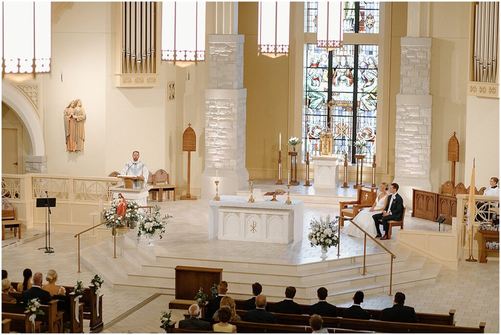 A bride and groom sit in chairs beside the altar at a Catholic wedding mass.