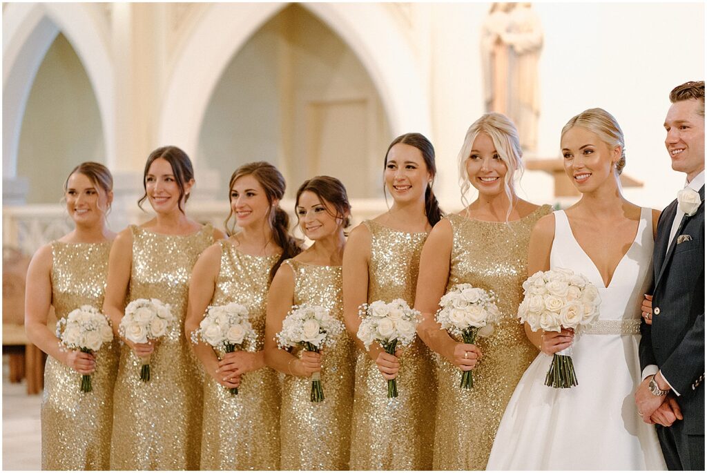 A bride poses besides a line of women in gold bridesmaid dresses.