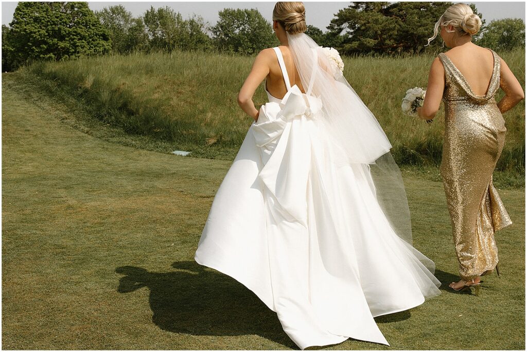 A bride and bridesmaid run through a field toward the Club at Lac La Belle.