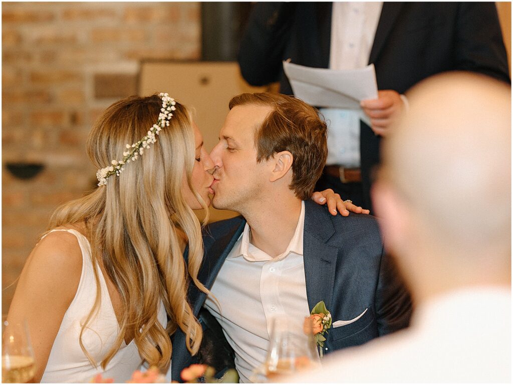 A bride and groom kiss at a dinner table in a candid wedding photo.