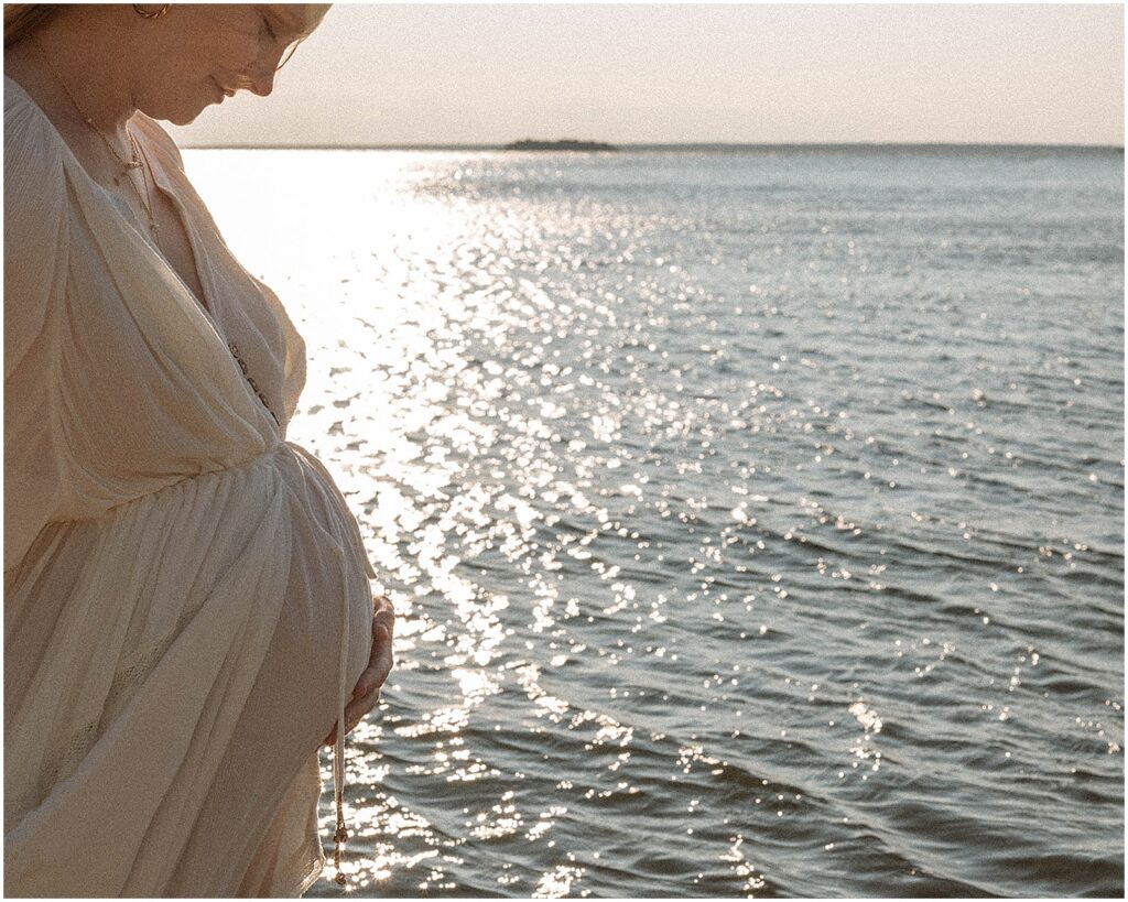 A woman looks down at her baby bump in front of a lake while she poses for a Door County Photographer.