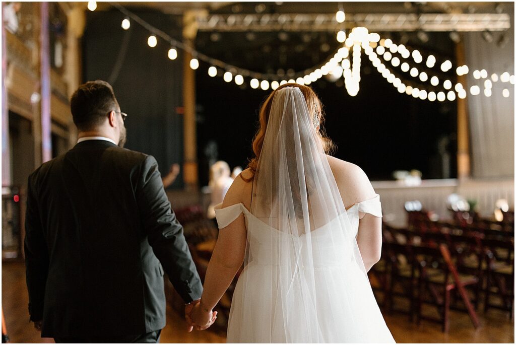 A bride and groom hold hands and look at lights strung over their ceremony venue.