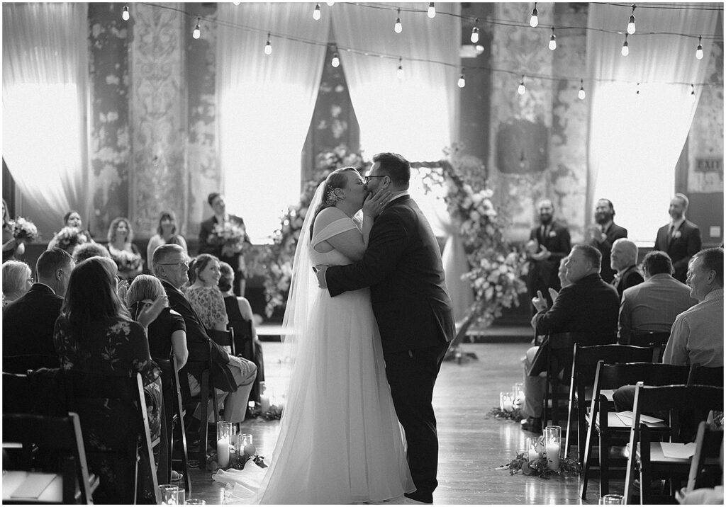 A bride and groom kiss at the end of the aisle during their recessional in a black and white wedding photo.