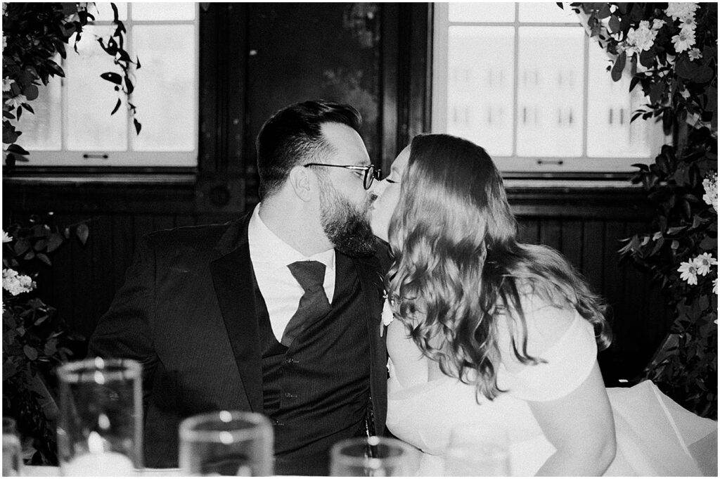 A bride and groom kiss at their reception table in a black and white wedding photo on film.