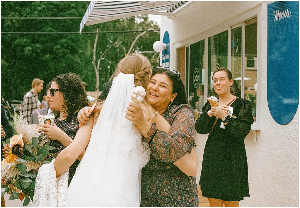 A wedding guest holding an ice cream cone hugs a bride outside a Milwaukee ice cream shop.