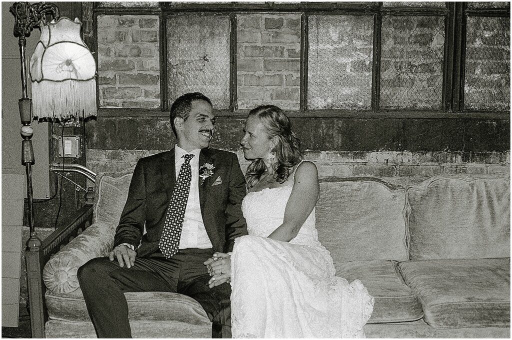 A bride and groom smile at each other on a vintage couch in a Milwaukee wedding venue.
