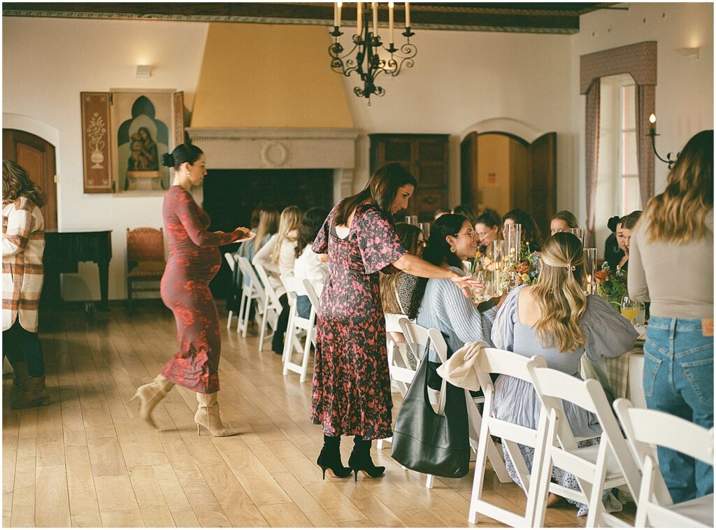 Two women walk around a table of baby shower guests in candid photography on film.
