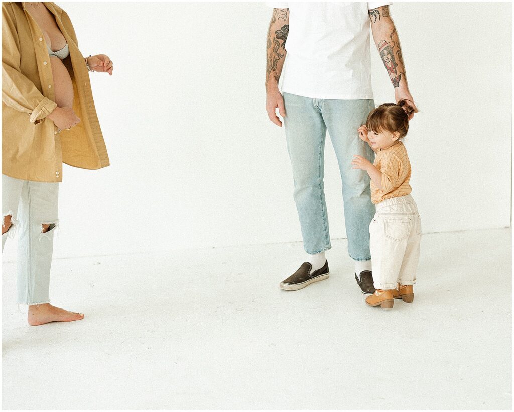 A toddler hugs her father's leg as her mother walks towards her.