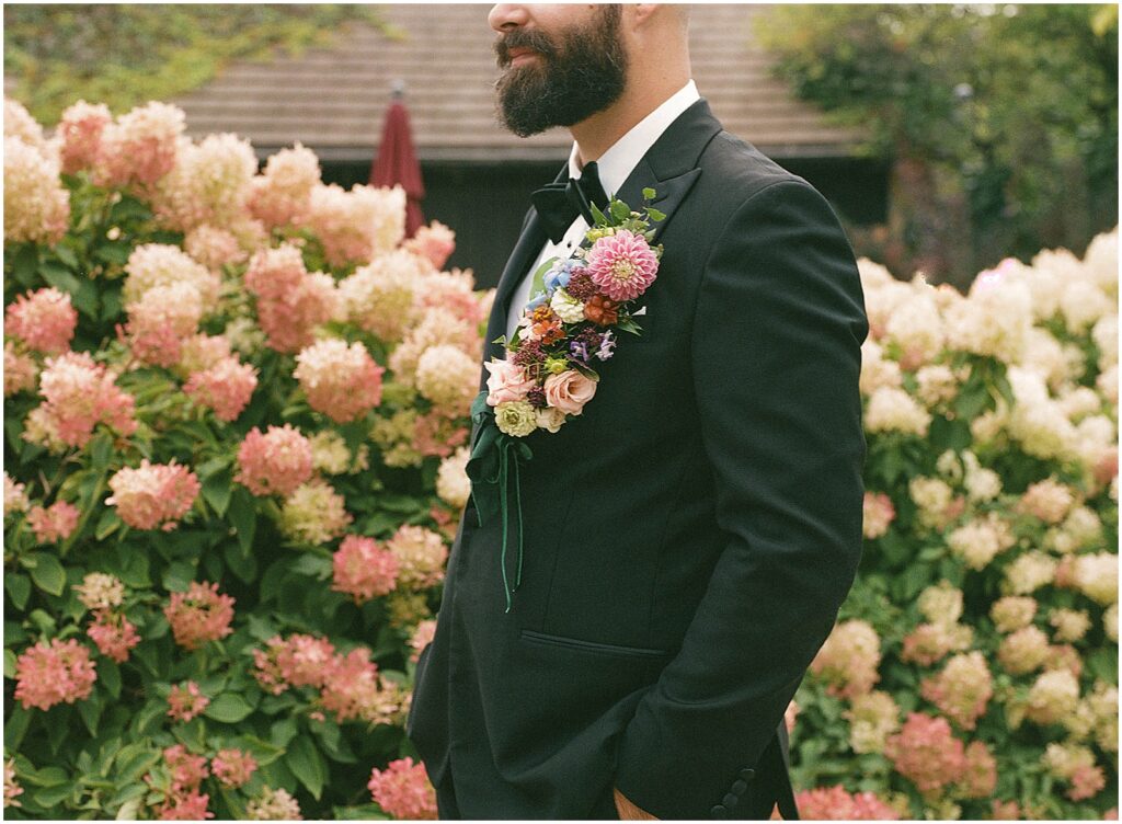 A groom with a floral boutonniere stands in front of a flowering bush at a garden wedding.