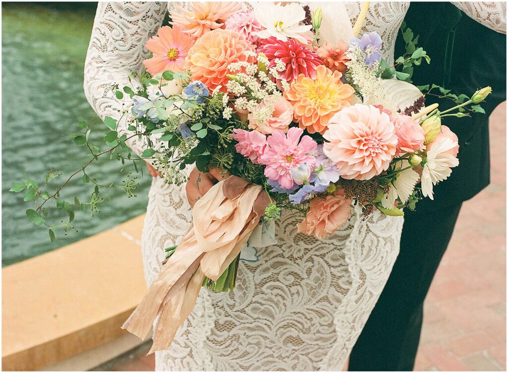 A bride holds a colorful wedding bouquet beside a fountain.