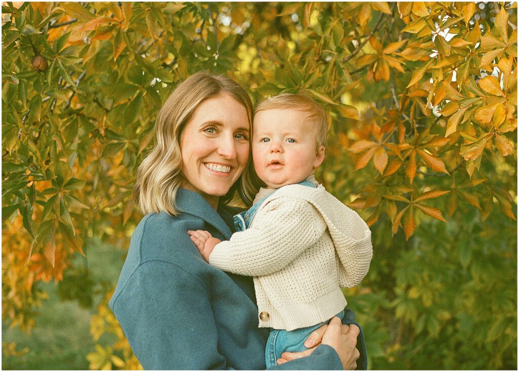 A woman poses with her baby in front of a tree with orange and green leaves.