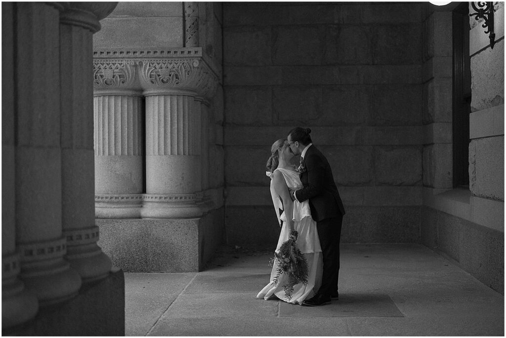 A bride and groom kiss between the pillars of a Milwaukee building in a black and white wedding photo.