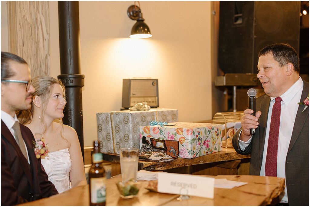 A bride and groom greet a guest beside the gifts table.