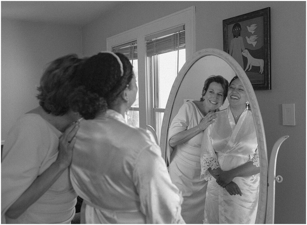 A bride's mother squeezes her shoulders as they smile in a mirror.