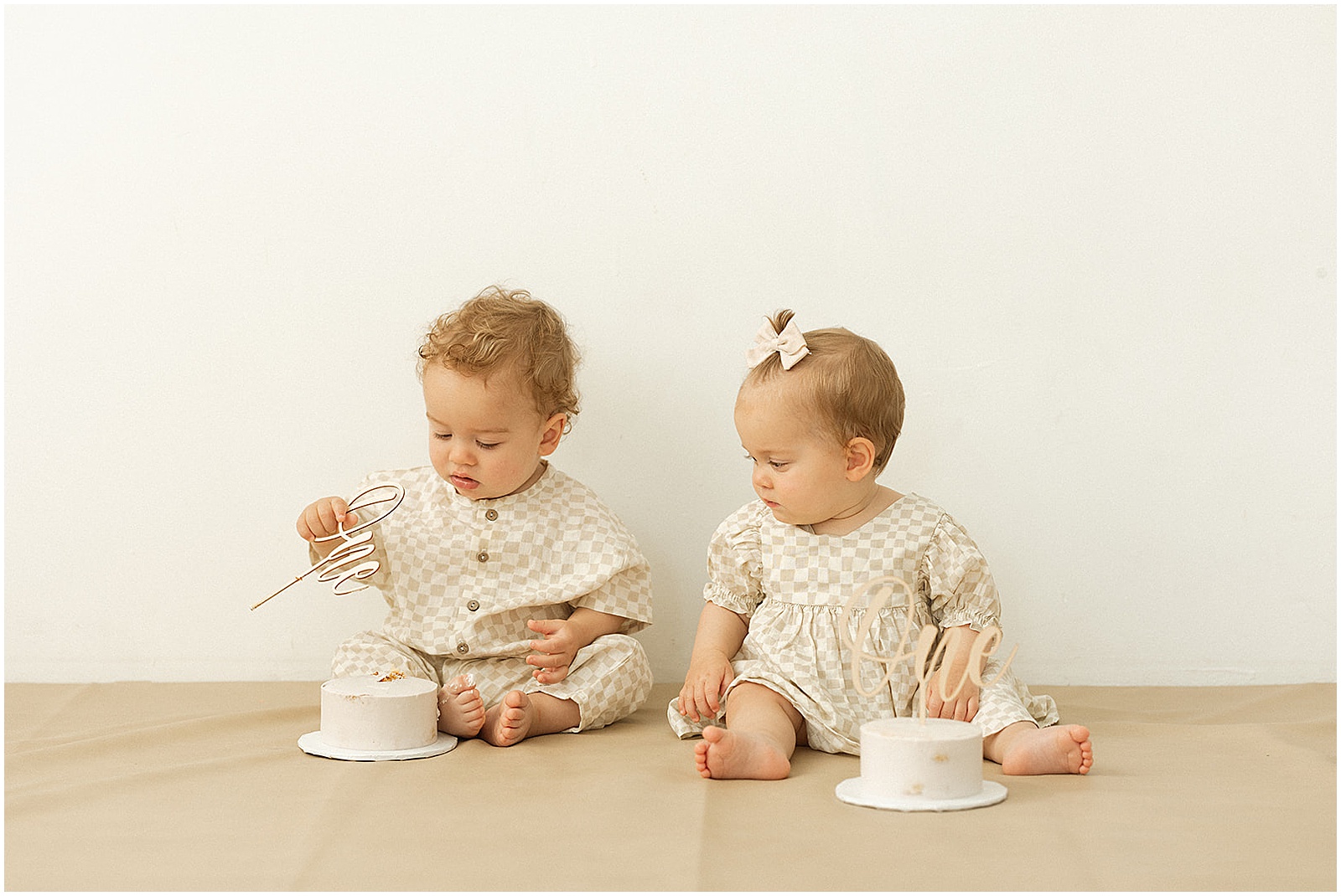 Twin babies sit next to each other looking at birthday cakes at a first birthday photoshoot.