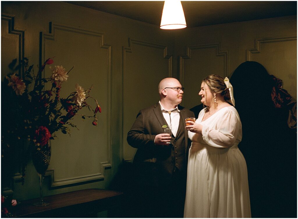 A bride and groom toast cocktails in a hallway on a film photo.