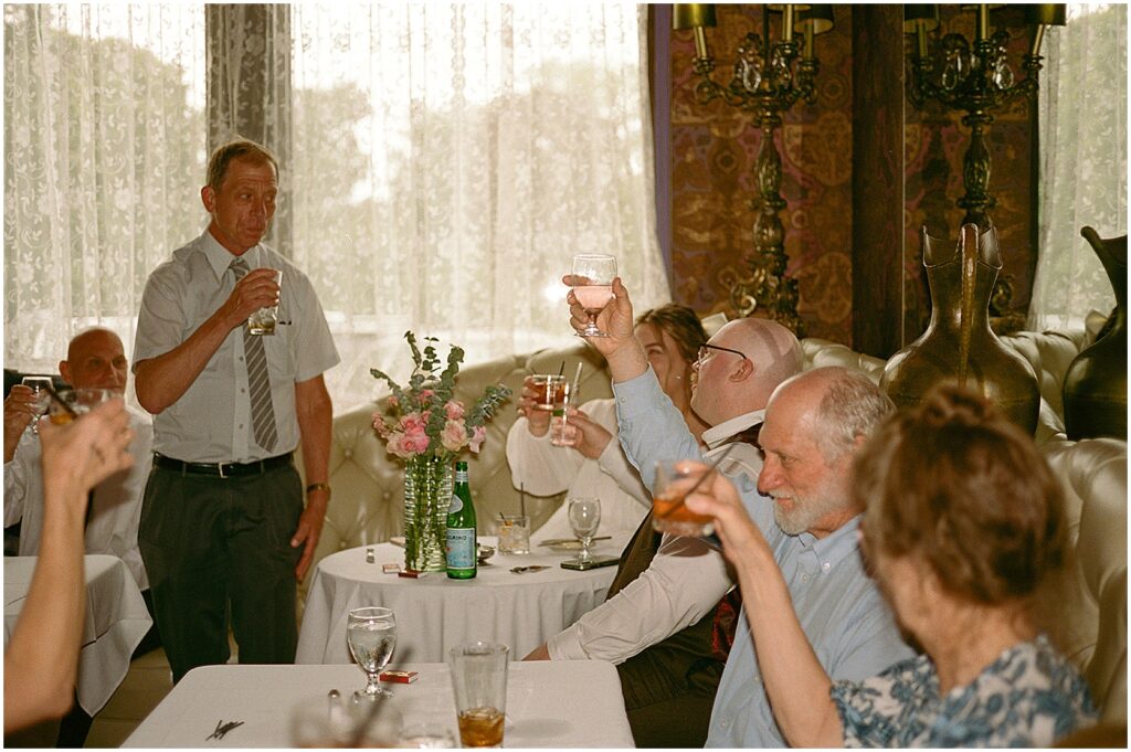 A man gives a wedding toast beside a window with lace curtains at the Hobnob Restaurant.