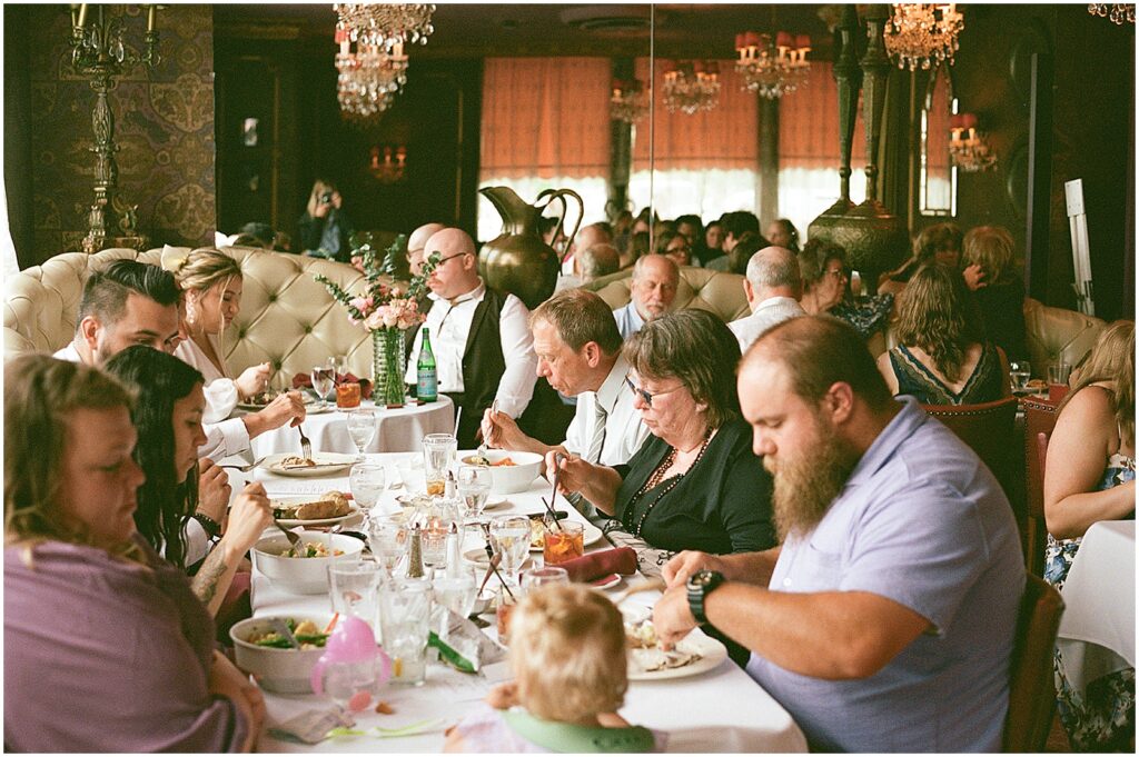 Wedding guests talk and eat dinner at long tables in a sunny room.