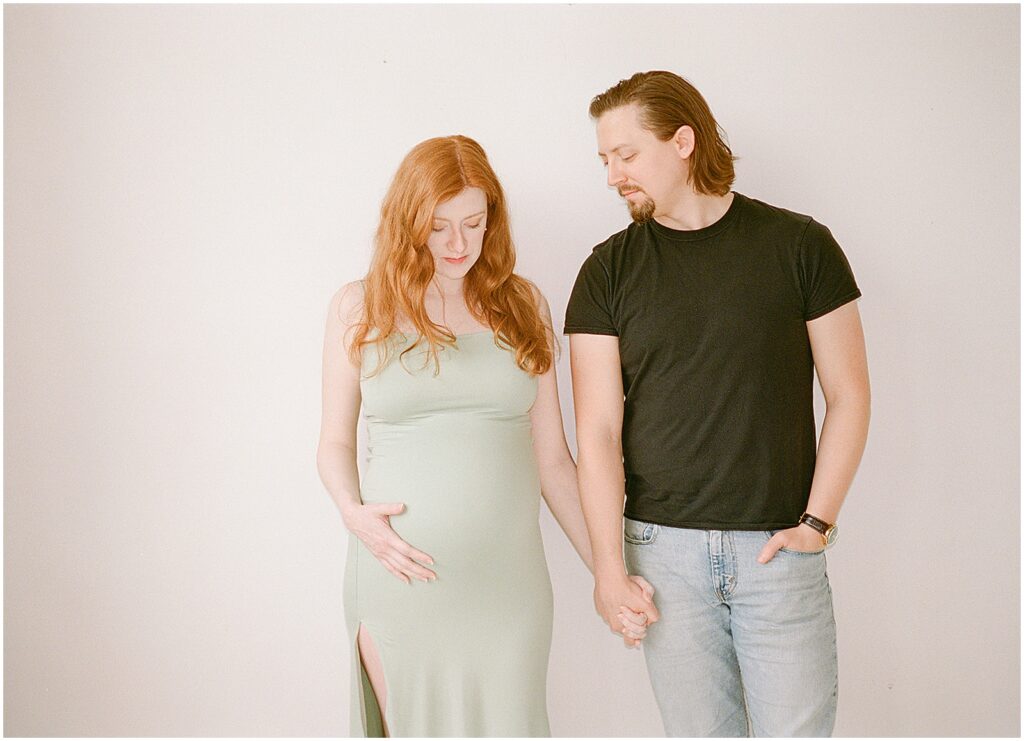 A man and woman hold hands in a Milwaukee photography studio and look at her pregnant belly.