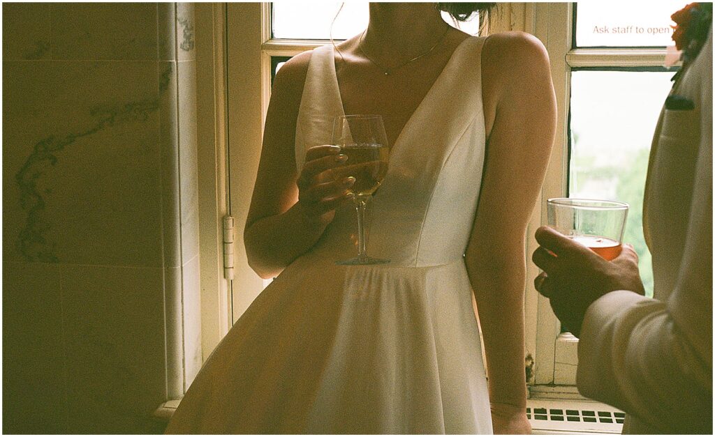 A bride and groom lean against a windowsill drinking glasses of wine.