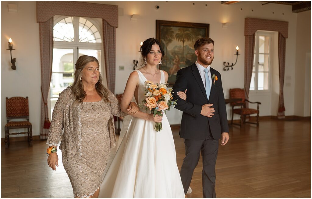 A bride's mother and brother take her arms to walk her up the aisle.