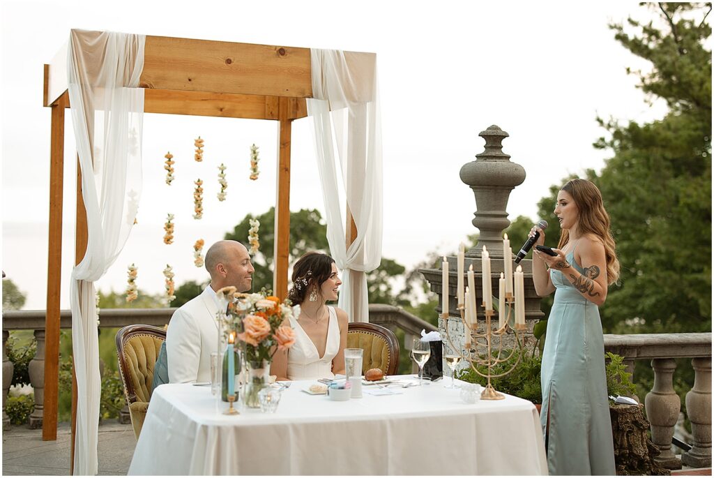 A bride and groom listen to a maid of honor's speech on a balcony at their Villa Terrace Milwaukee wedding.