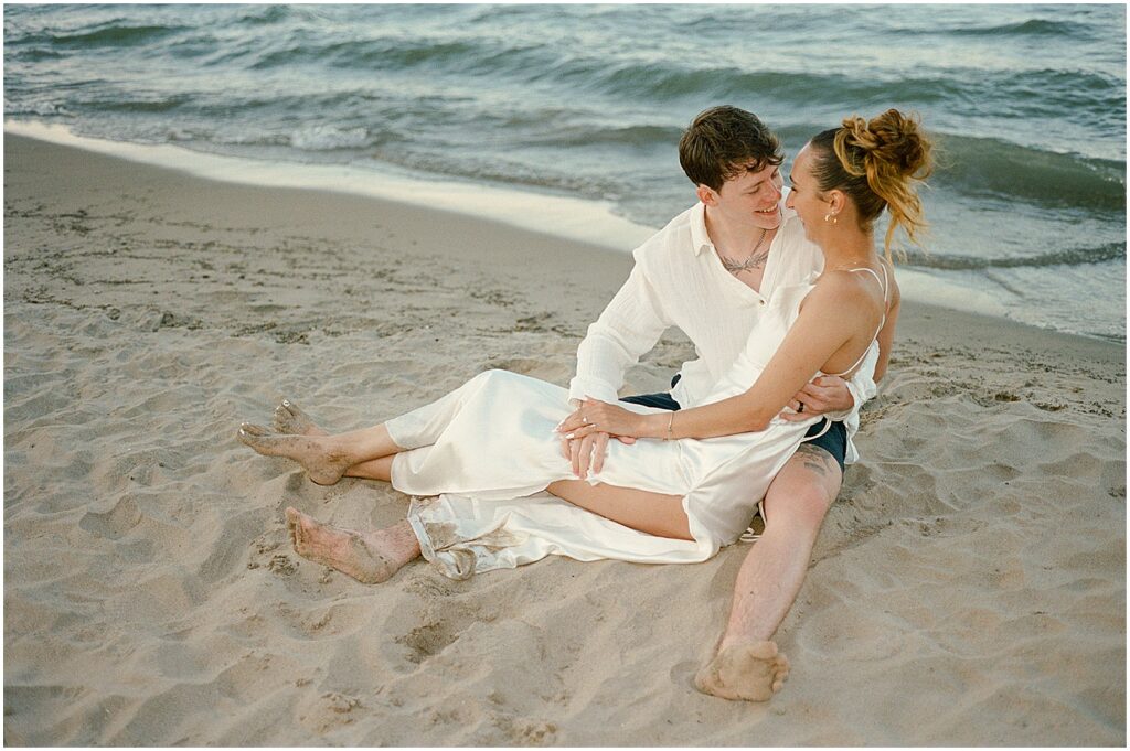 A bride sits in a groom's lap in the sand beside Lake Michigan.