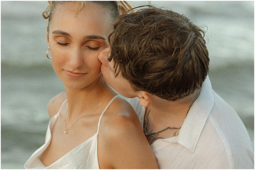 A groom kisses a bride's cheek on a beach in Milwaukee.