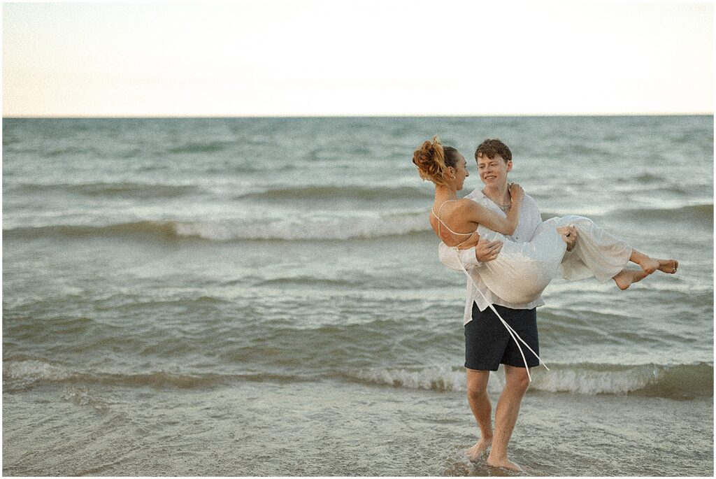 A groom carries a bride across a beach towards a Milwaukee elopement photographer.