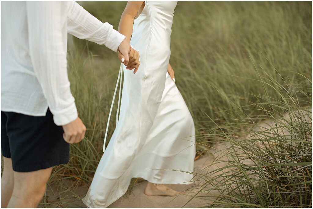 A bride holds a groom's hand on a sandy path through Atwater Beach.