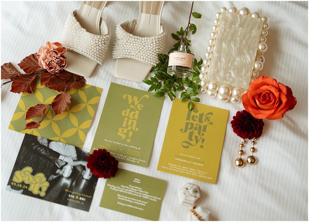 A green wedding invitation suite lays on a hotel bed with shoes, a purse, and florals.