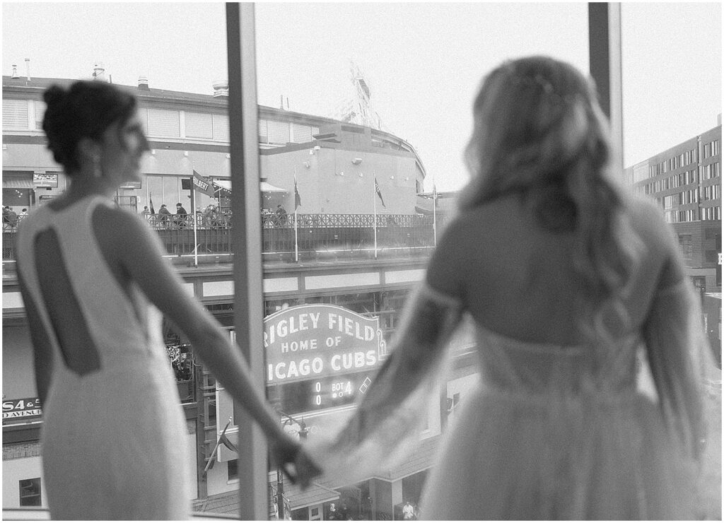 Two brides hold hands in a hotel room overlooking Wrigley Field.