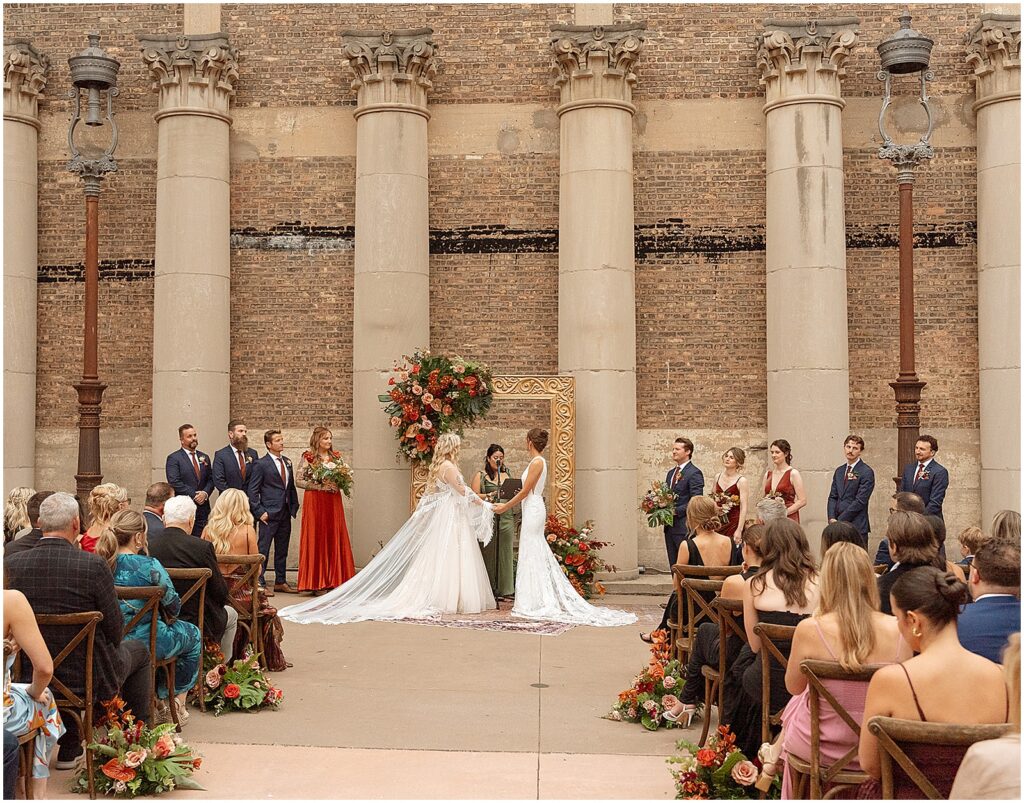 Two brides hold hands during a courtyard ceremony at their Artifact Events wedding.