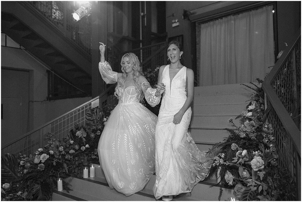 Two brides enter their Artifact Events wedding reception from a staircase lined with flowers.